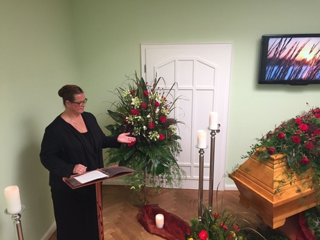 Birgit hält Rede in Raum mit Kerzen, Blumengestecken und geschmücktem Sarg